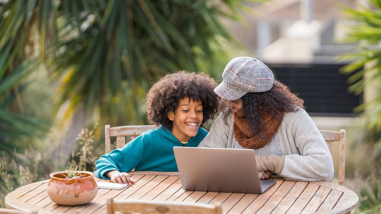 A mother works with her young son on a laptop at a table outside.