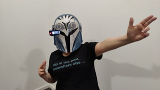 Person doing finger guns whilst wearing the Black Series Bo-Katan helmet with the heads-up display down and lit up. They are wearing a black t-shirt with light blue text that reads “Way in the past, somewhere else…”.