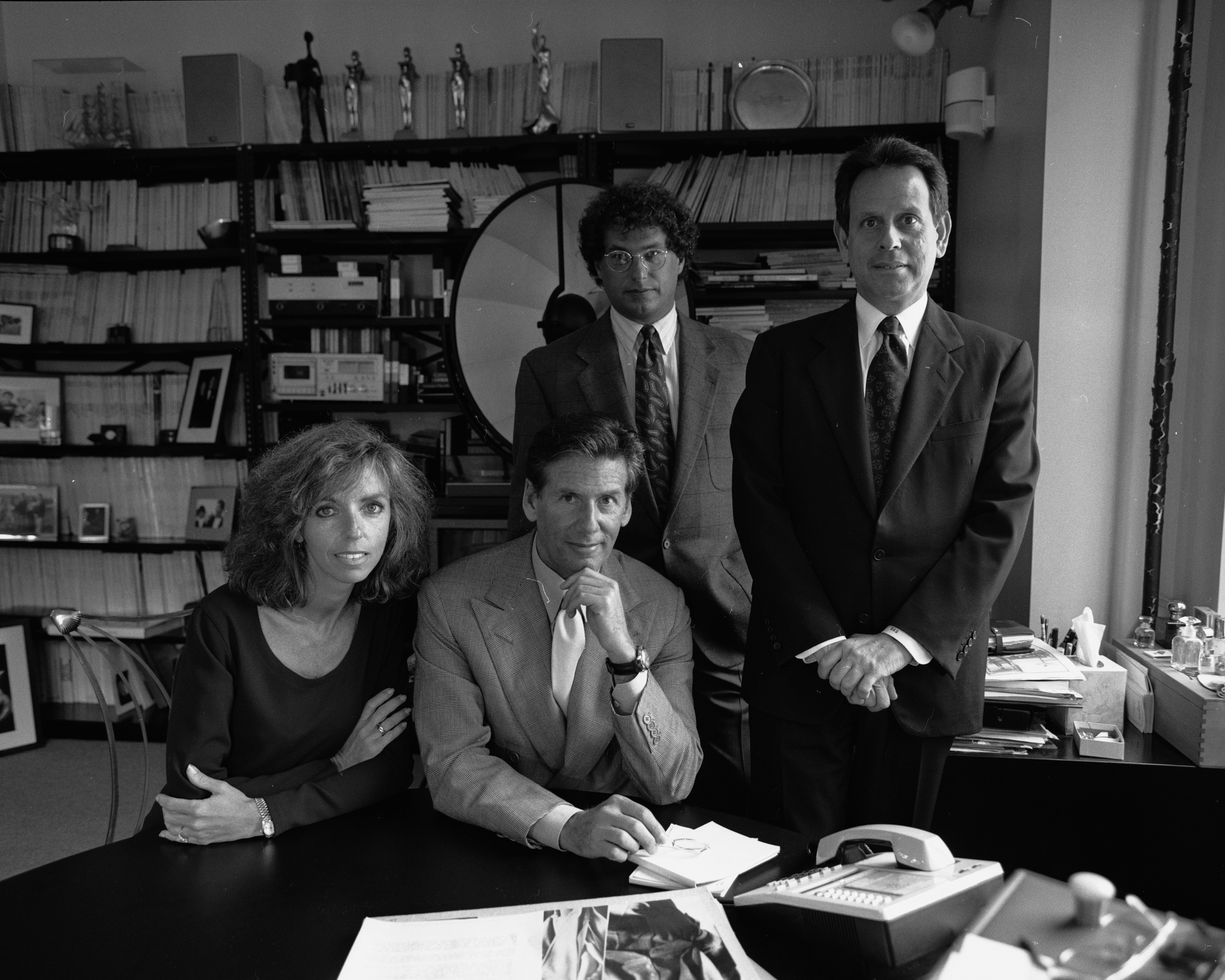 A group of beautifully dressed people in professional attire sit behind a desk in a books, awards, pictrues, and documents-filled office while posing for the camera in a black and white picture.
