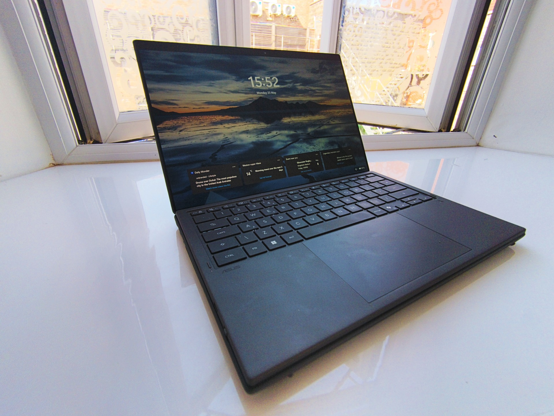 Side angle view of the ASUS Zenbook Duo in traditional laptop mode, displaying the Windows lock screen showing the time 15:52 against a mountain landscape background next to an open window.