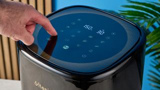 a photograph of a finger pressing the touchscreen icons of a large black 5.3 quart capacity air fryer with black touchscreen and rose gold embellishments sits upon a brown table with a blue background