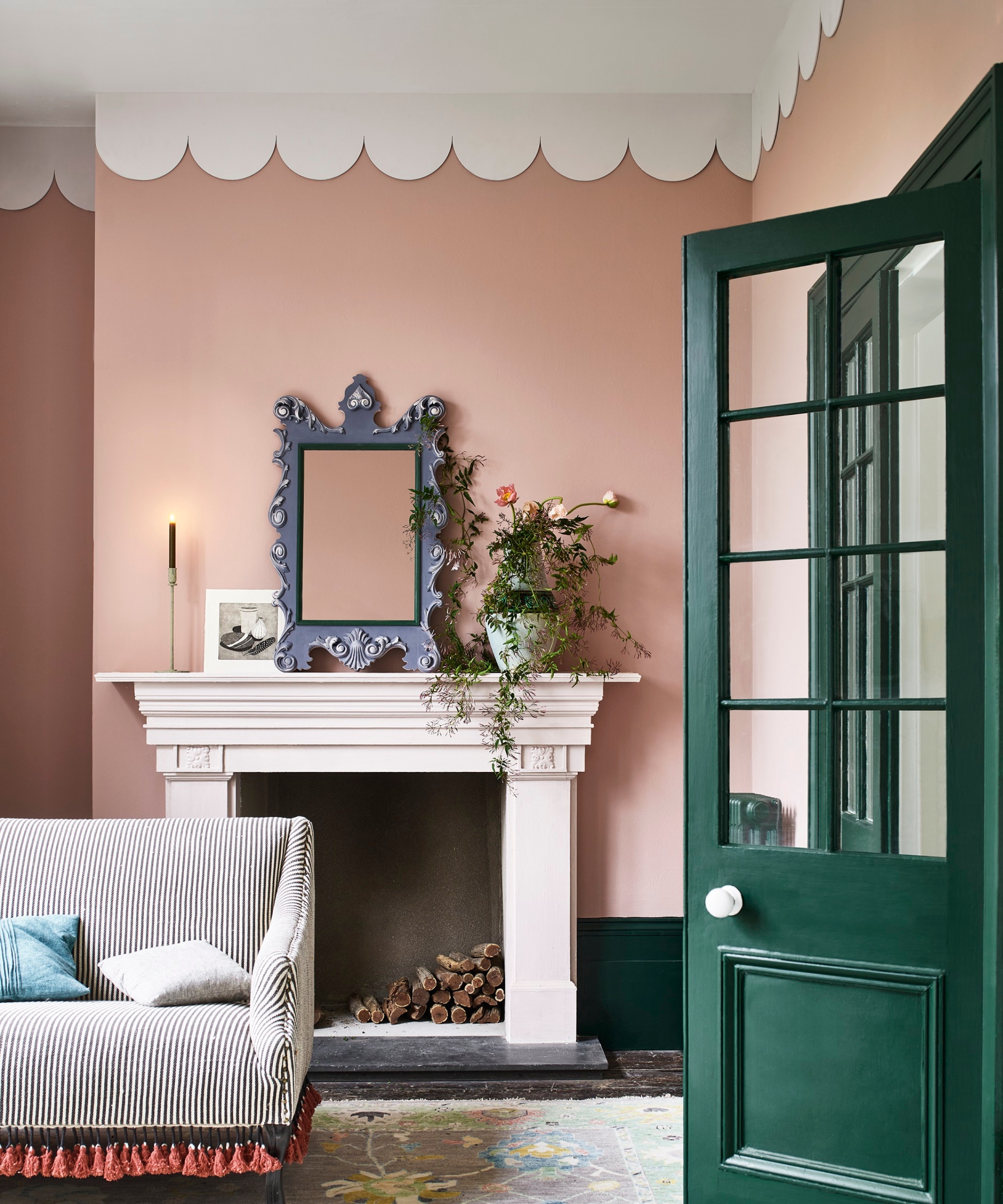 Pink room with scalloped ceiling border, ornate mirror over a fireplace, striped sofa, and a green door.