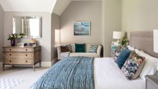 A neutral bedroom with a large double bed with white and blue bedding, cushions, and matching bedside tables with white lamps. Behind is a cream colored couch with cushions beneath a framed artwork, and to the right is a brown chest of drawers with a vase of flowers on top beneath a large rectangular mirror.