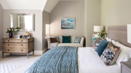 A neutral bedroom with a large double bed with white and blue bedding, cushions, and matching bedside tables with white lamps. Behind is a cream colored couch with cushions beneath a framed artwork, and to the right is a brown chest of drawers with a vase of flowers on top beneath a large rectangular mirror.