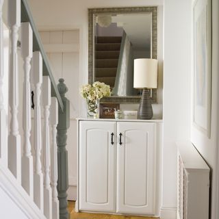 White and green stair case leading to wooden floor and white cupboard with black and white lamp and glass vase with white flowers