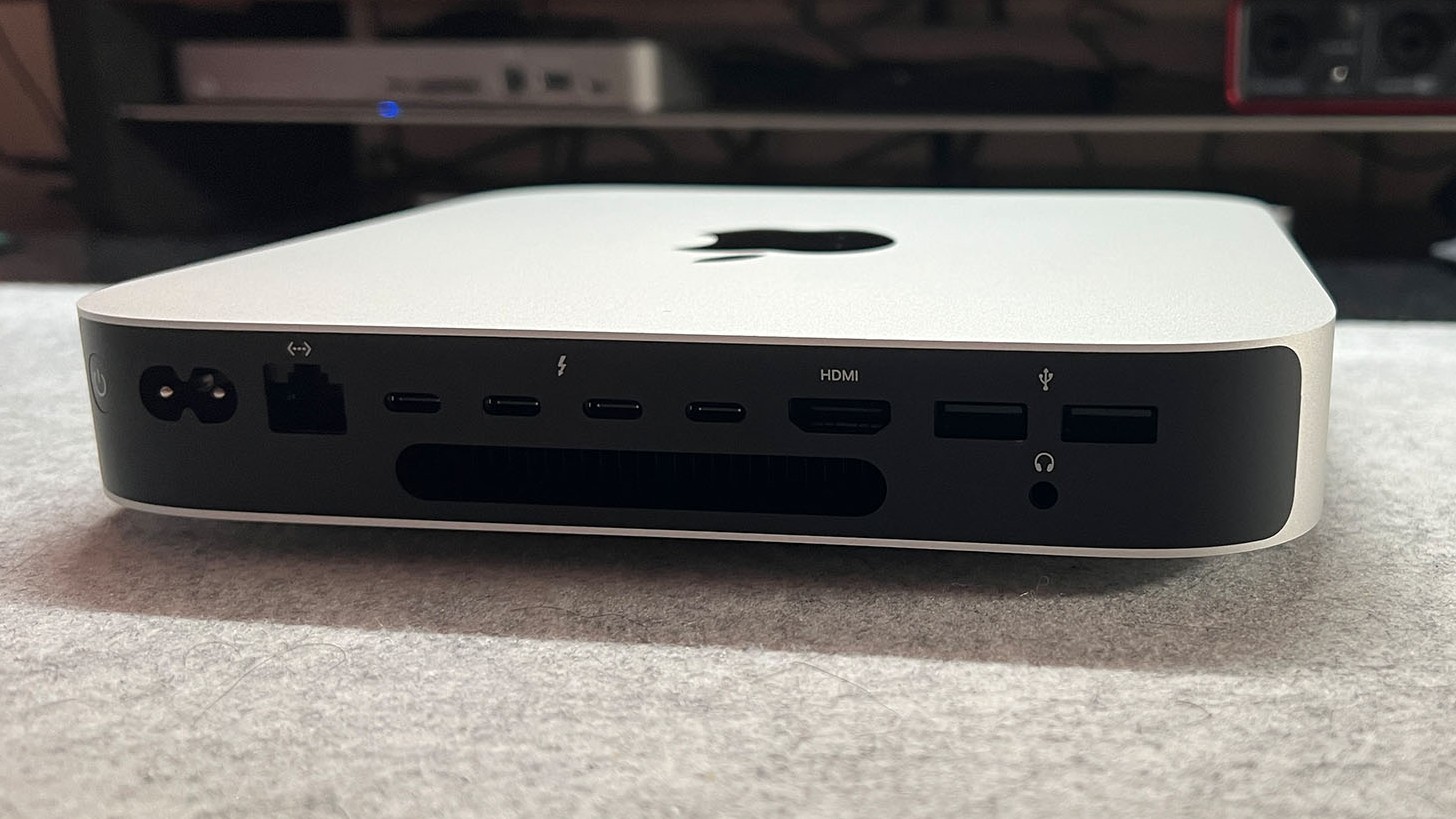 The Mac Mini Pro M2 is mini pc. It's a thin, square shape with rounded corners (20 x 20 x 3.5 cm). It's silver in color and has the Apple logo in the center of it (a black apple with a bite taken out of it). Here we see a close up of one edge – it's black and has several ports/inputs, such as a headphone jack.