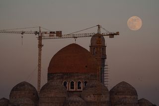 the moon rises over a mosque with cranes