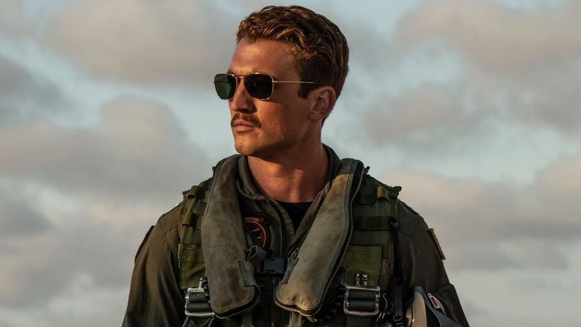 Miles Teller looks off to the side wearing full flight gear and aviators in Top Gun: Maverick.