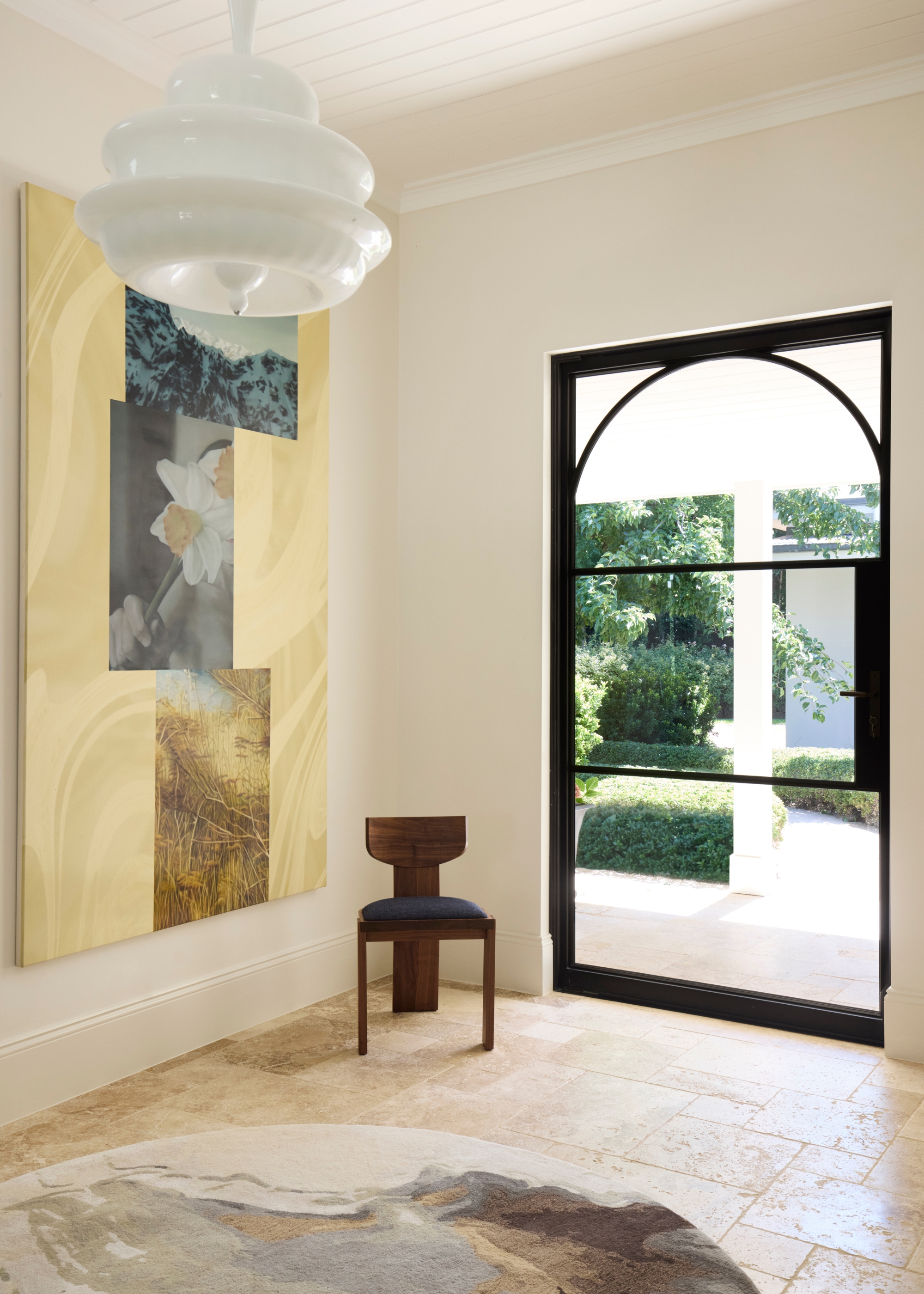 an entryway with a black-framed glass front door framed with an internal arch with a circular rug in front, a large piece of artwork on the wall, a white circular ceiling light and a modern chair in the corner of the space by the door