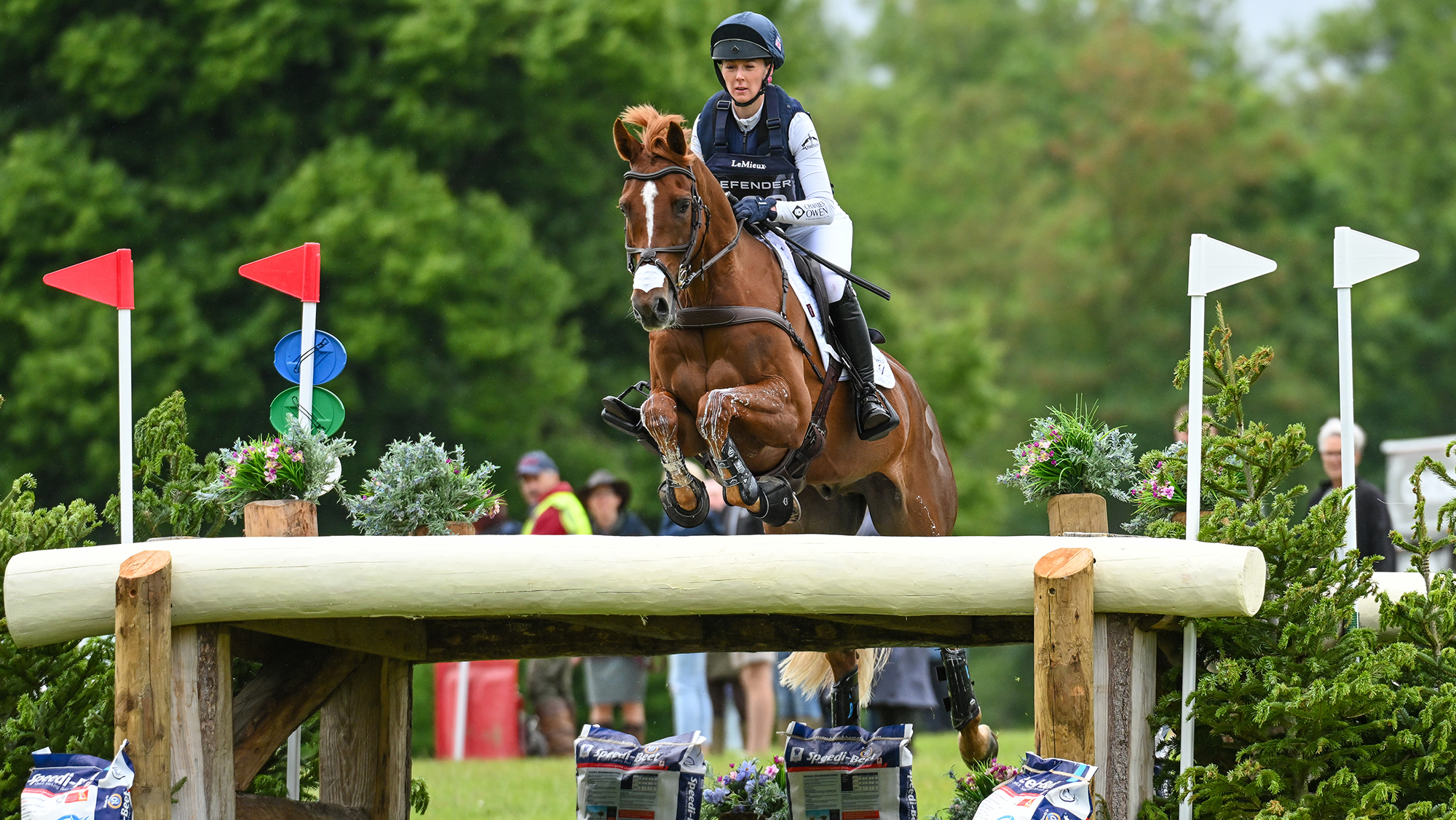 Yasmin Ingham, a rider in a white top with a blue body protector over it, rides chestnut horse Goliath Du Loir over a table fence at Bramham 2026