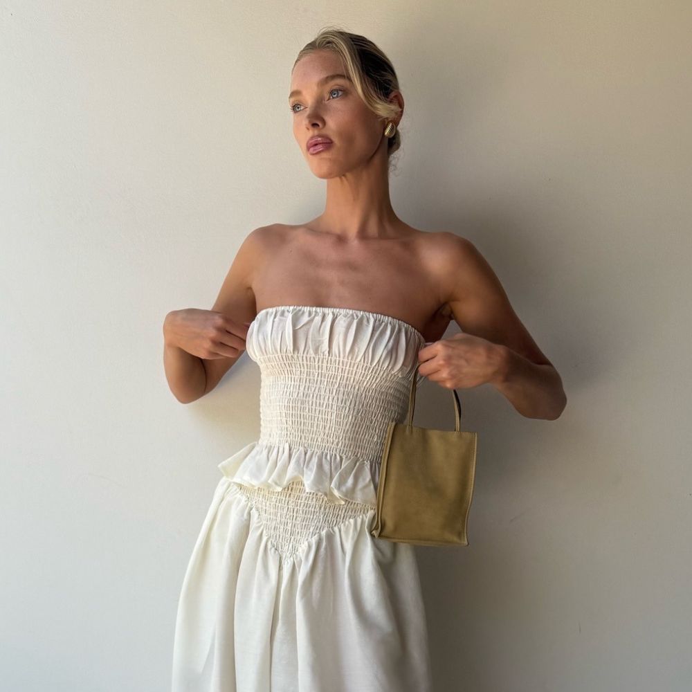 Elsa Hosk stands inside against a white wall wearing a white strapless two-piece with gold woven flats from Alaia.