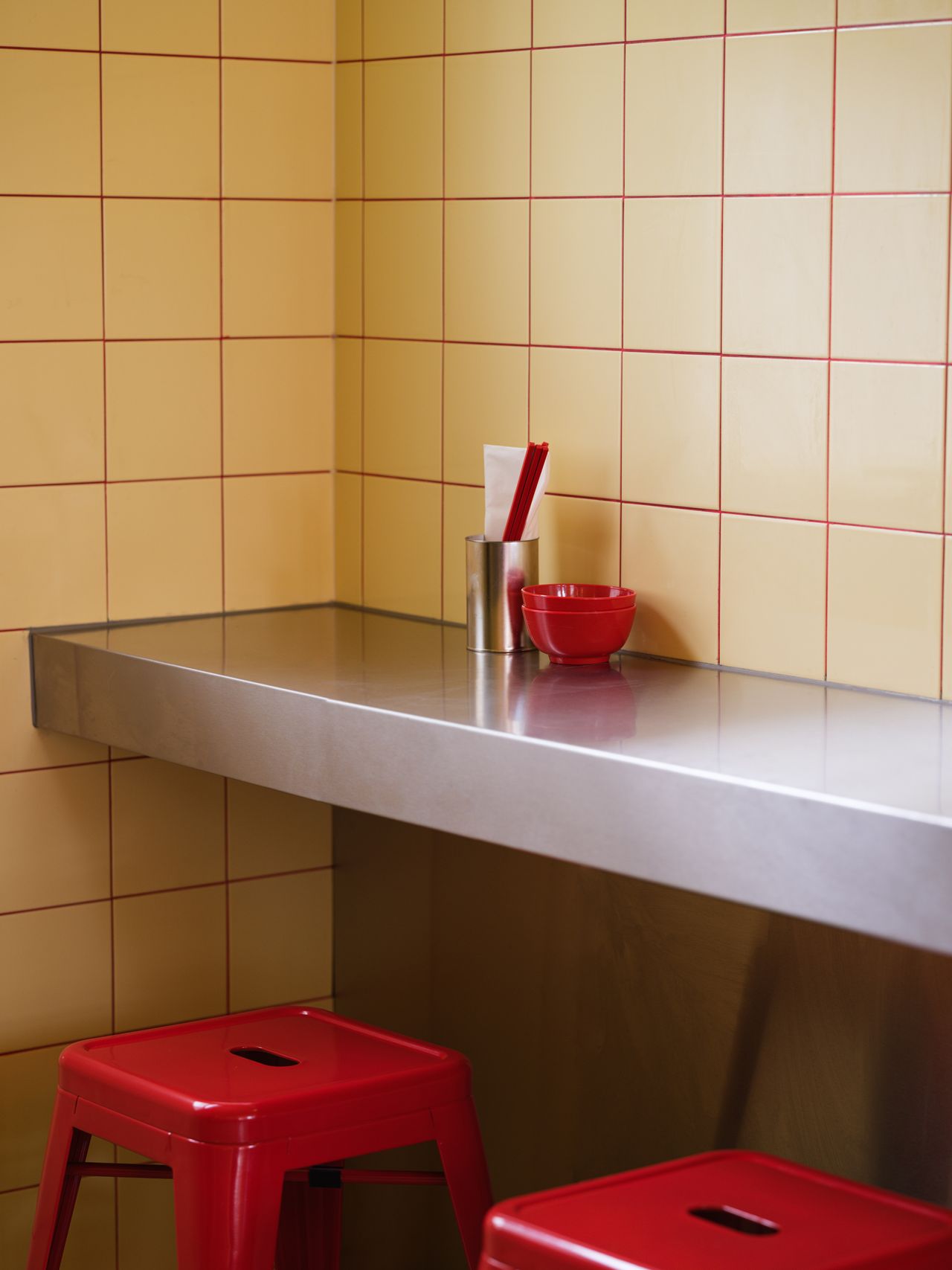 A stainless steel matt counter, paired with lacquered red stools and matching Asian tableware in red and silver, is positioned against a tiled, buttery yellow wall with red detailing.