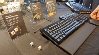 Be Quiet's modular Dark Mount mechanical gaming keyboard at an announcement press event. A hand reaches into frame, demonstrating the detachable modular wrist rests, alongside the detachable number pad and media dock. Be Quiet's orange silent switches are scattered nearby.