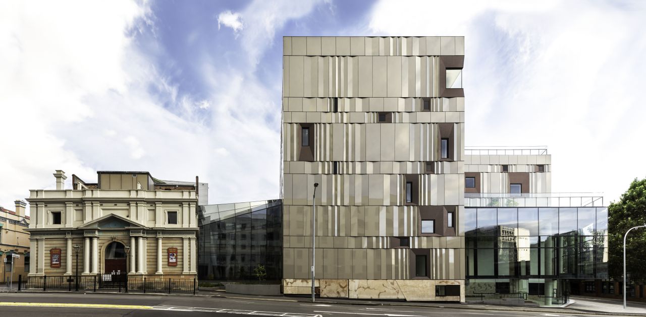 Daytime image of the dramatic exterior of the Hedberg building from the street side in Tasmania, road at the front of the building, Trees to the right, stone building to the right, street light, blue cloudy day sky