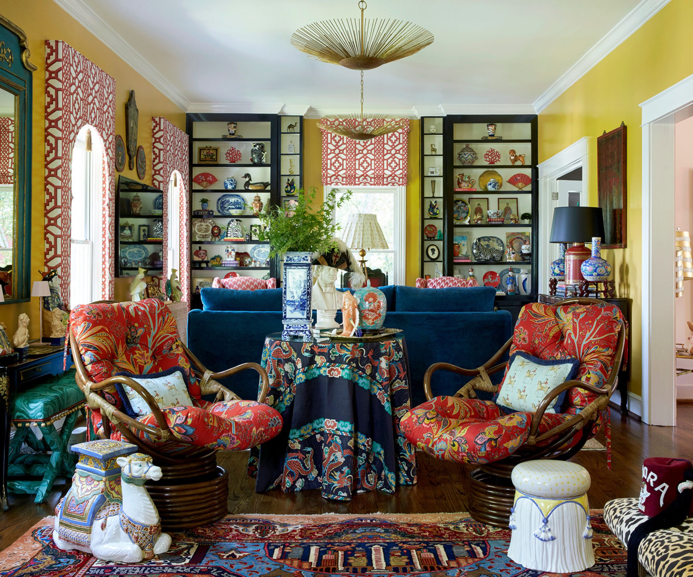 maximalist living room with yellow walls, bookcases filled with antiques, large blue couch, red upholstered antique chairs, ceramic stool and leopard bench
