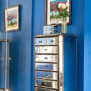 a mirrored skinny tall chest of drawers with gold trim, in front of a blue wall, with a mirrored cupboard next to it