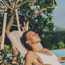 A woman relaxing on a wooden deck chair by a pool and some palm trees.