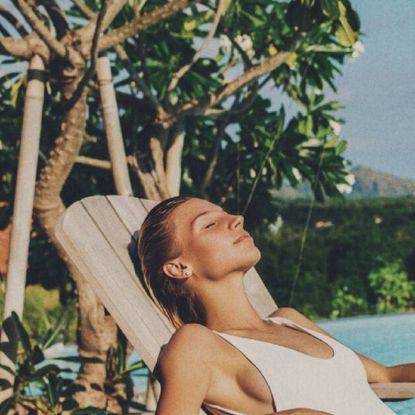 A woman relaxing on a wooden deck chair by a pool and some palm trees.