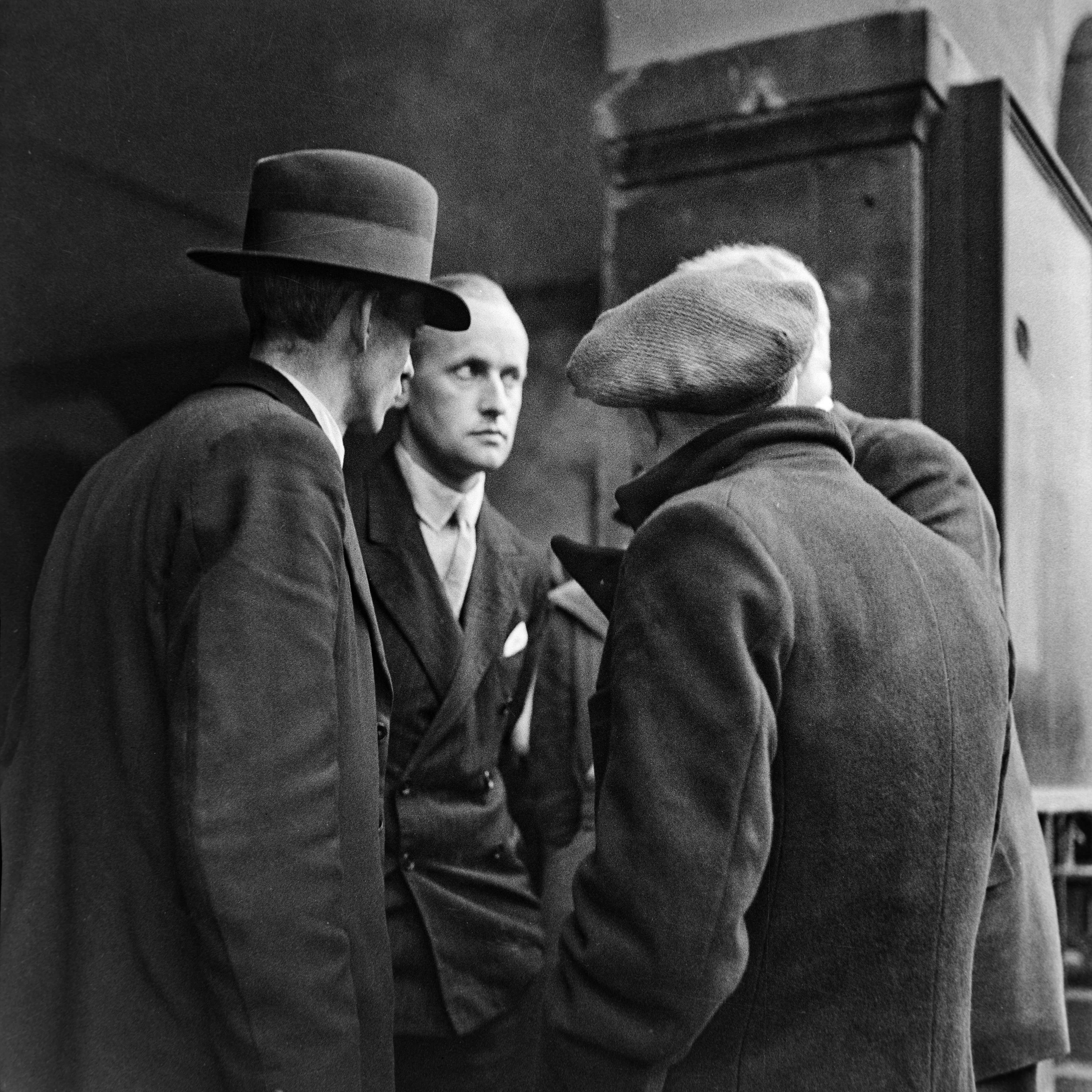 Three men in heavy overcoats stand in close conversation outside a large stone building. The central figure faces the camera directly with a tense, watchful expression. The image is tightly framed, giving it an intimate and slightly conspiratorial quality.