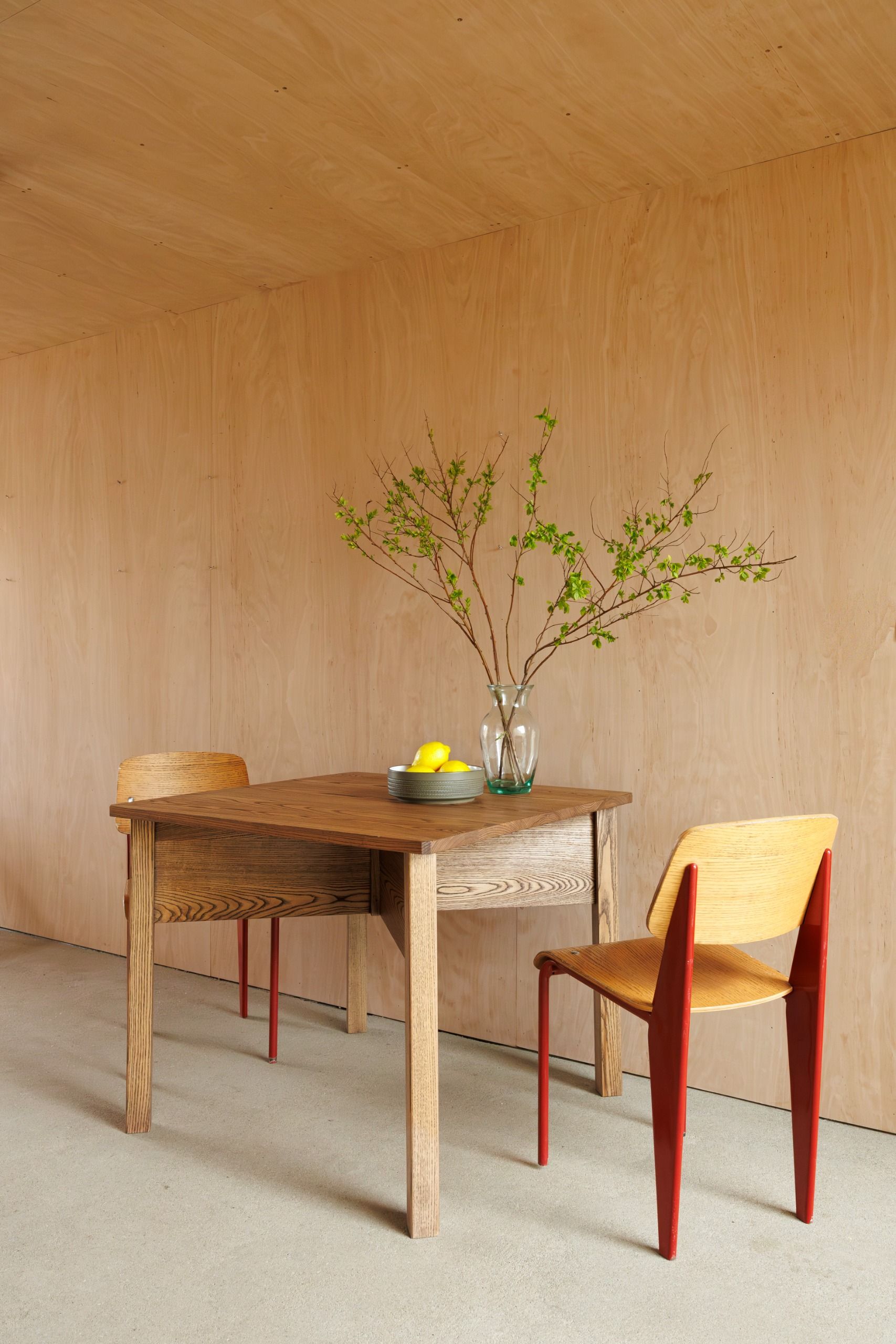 A wood-paneled living room with a central, square table, red-painted wooden chairs, a transparent glass vase, and a fruit bowl filled with oranges, looking out to greenery.