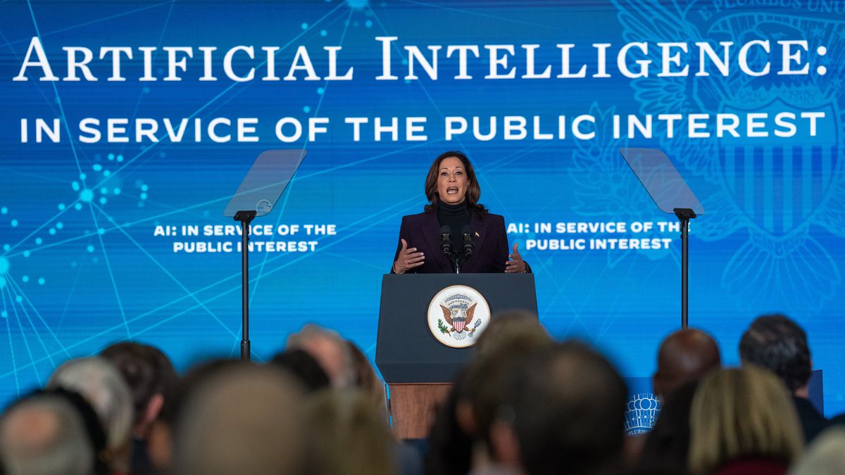 Kamala Harris, vice president of the United States of America, delivering an address on AI policy at the US Embassy in London. Harris is stood onstage behind a podium, with a large blue background behind her bearing the US presidential seal and the words &quot;Artificial intelligence: in service of the public interest&quot;.