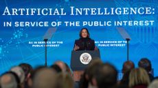 Kamala Harris, vice president of the United States of America, delivering an address on AI policy at the US Embassy in London. Harris is stood onstage behind a podium, with a large blue background behind her bearing the US presidential seal and the words "Artificial intelligence: in service of the public interest".