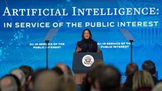 Kamala Harris, vice president of the United States of America, delivering an address on AI policy at the US Embassy in London. Harris is stood onstage behind a podium, with a large blue background behind her bearing the US presidential seal and the words "Artificial intelligence: in service of the public interest".