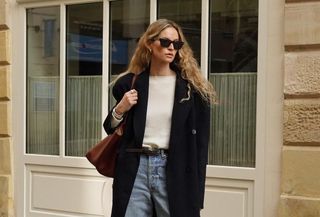 @anoukyve wearing an elegant fall outfit consisting of a black wool coat, cream sweater, jeans, and suede handbag.