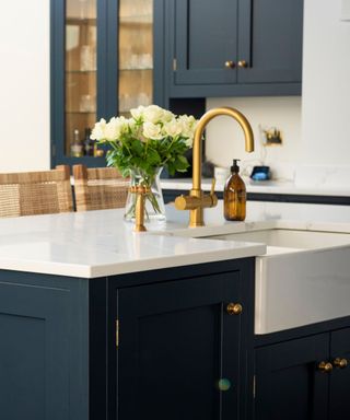 A kitchen island with a white surface, a brushed gold faucet, a glass vase of white roses and a brown soap dispenser, and navy blue cabinets with gold handles below it and on the walls behind it