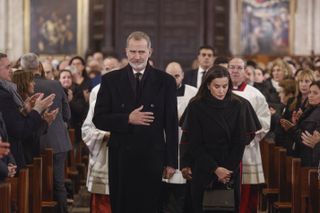 King Felipe and Queen Letizia wearing dark coats walking down the aisle of a church as people clap