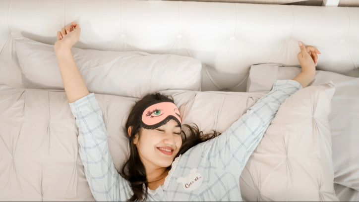 A woman wearing a pink sleep eye mask and silk pyjamas stretching her arms in bed waking up happy with her sleep quality.