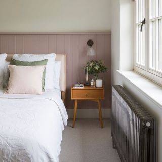 White painted bedroom with pink panelling behind the bed with white bedding on it, and a radiator underneath the window