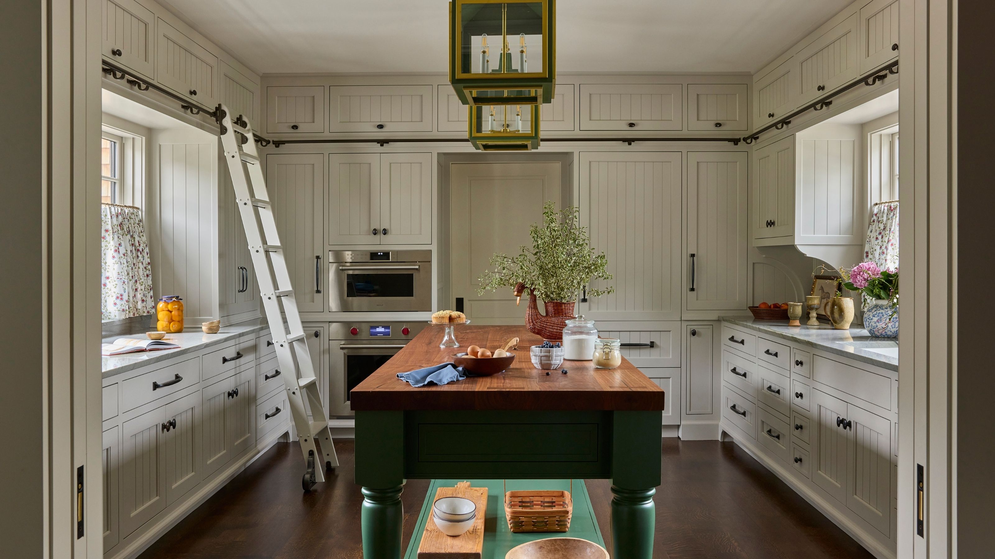 Off-white country kitchen with extensive cabinetry, integrated appliances, a rolling library ladder, and a central dark green kitchen island topped with a butcher-block counter.