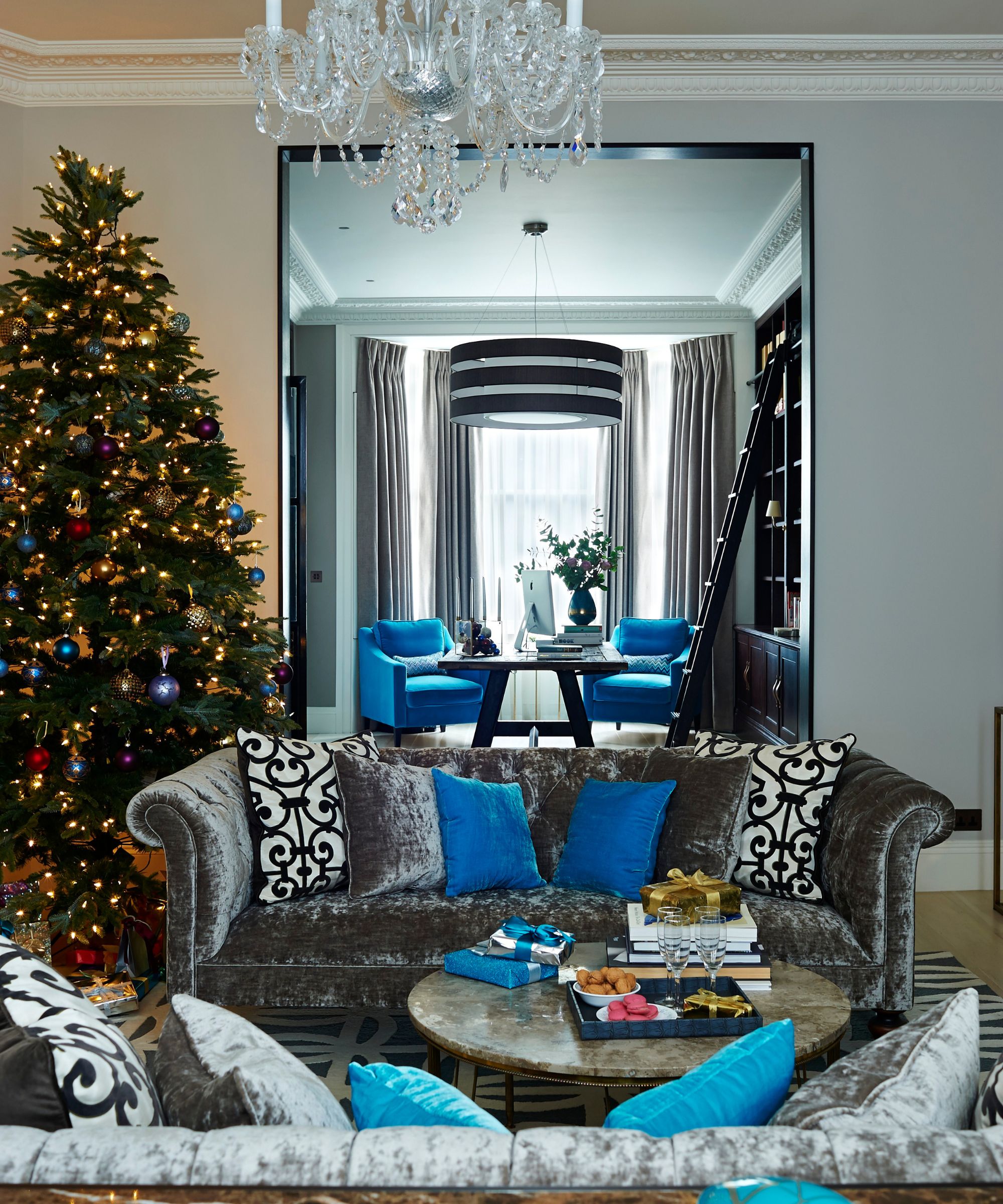 A Christmas living room with two grey crushed velvet couches, patterned cushions, a circular marble coffee table between them with presents, books and a tray of food and drinks on it. To the left is a large decorated Christmas tree and behind is a living room through an open doorway.