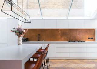 a white kitchen with a brass splashbacks. There is also a kitchen island with flowers on top, next to the island are three bar stools.