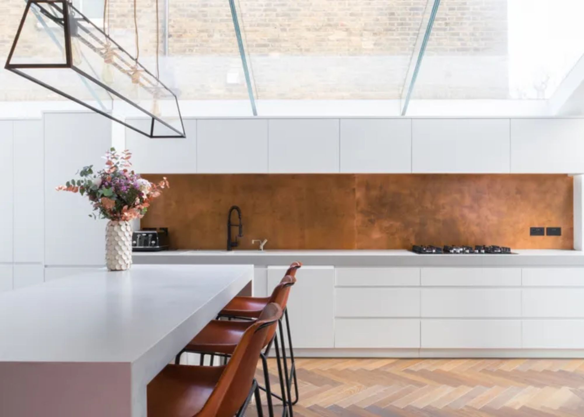 a white kitchen with a brass splashbacks. There is also a kitchen island with flowers on top, next to the island are three bar stools.