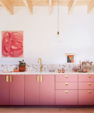 Pink kitchen cabinets with gold handles, pink paintings on the walls and a terrazzo-style splashback