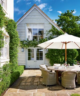 A curved wooden table and wicker chairs demonstrating patio furniture ideas in a small, country-stile patio with climbing plants.