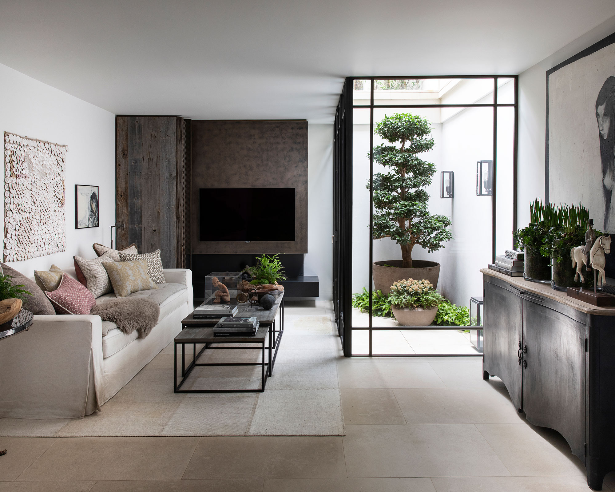 A contemporary living room with a neutral sofa, nested coffee tables, and a TV accent wall, featuring a glass-enclosed indoor atrium that holds a large, manicured bonsai-style tree.