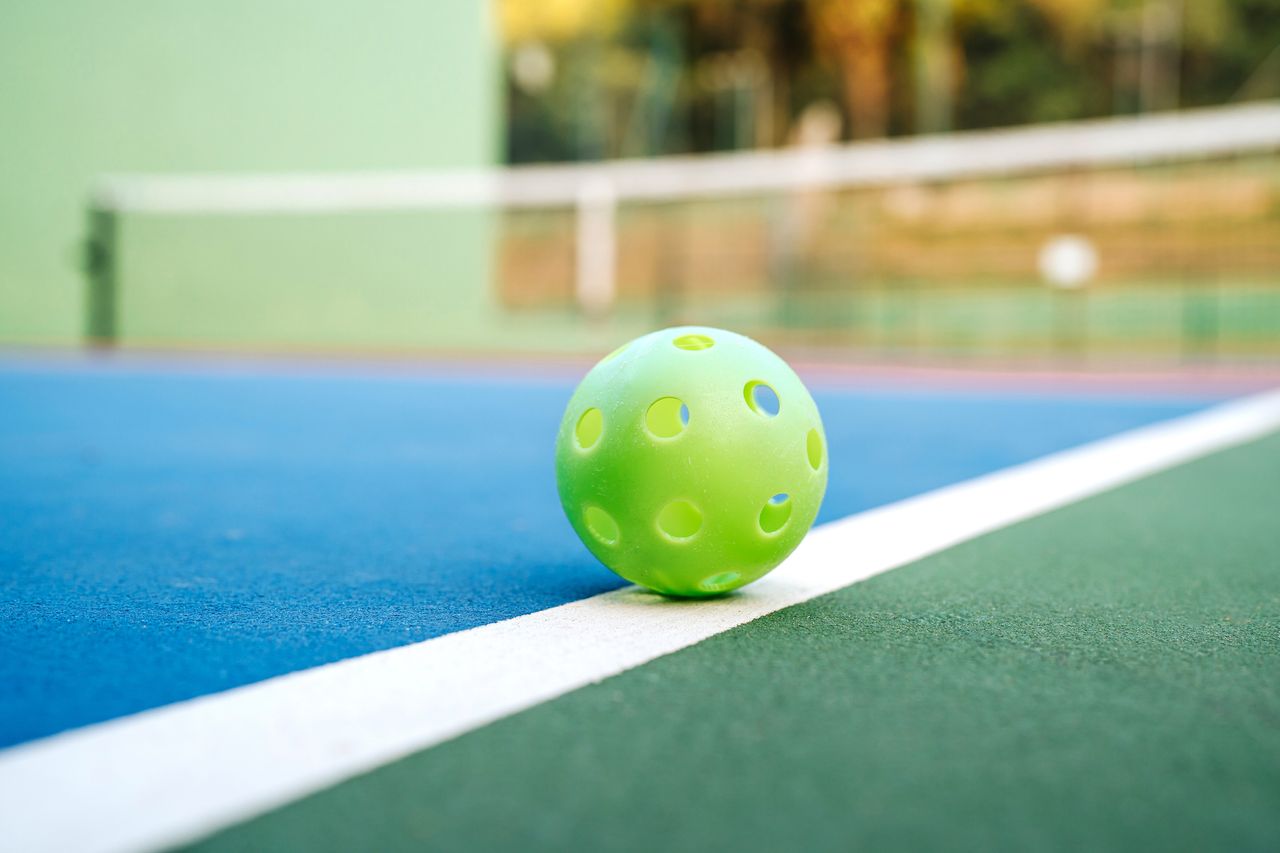 green-yellow perforated ball on a pickleball court with a net and vegetation in the background