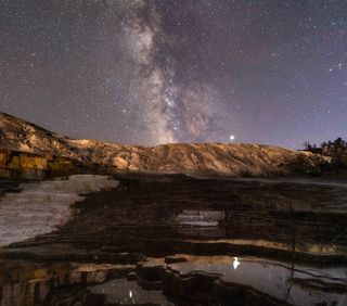 The Milky Way shimmers over Yellowstone National Park in this starry night-sky photo by Chirag Upreti. This panorama combines 10 frames captured from the "Mound and Jupiter Terrace" at Mammoth Hot Springs, and it features an excellent view of the planet Jupiter, shining brightly to the right of the Milky Way while reflecting off the surface of the water below. Saturn is also visible in the photo, shining to the left of the Milky Way, directly above the asterism known as the Teapot.
