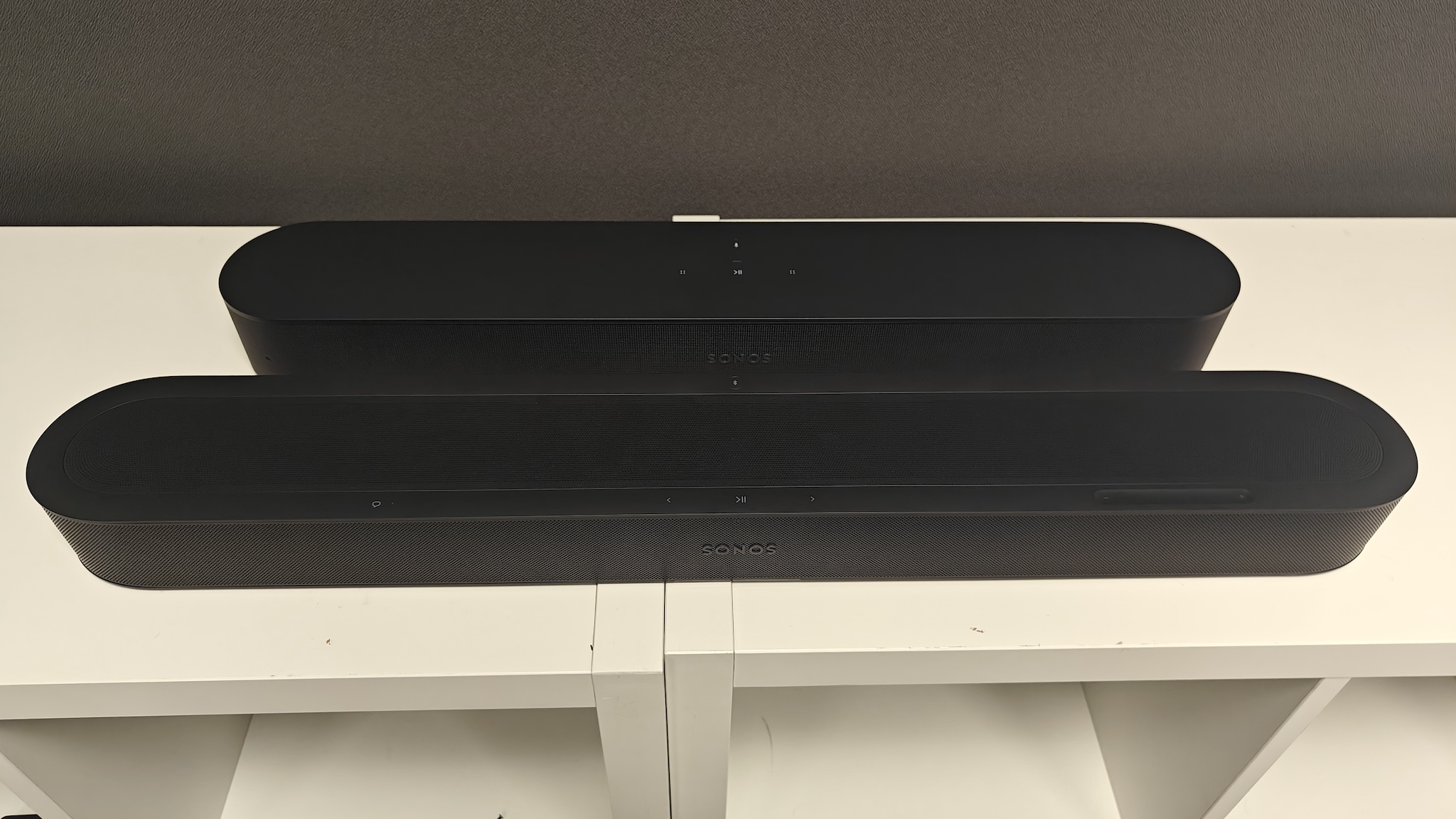 A black Sonos Beam Ultra and Beam (Gen 2) next to each other on a white TV cabinet, shot straight-on and from slightly above.