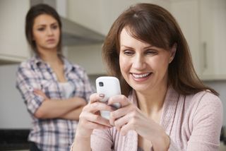 Mother texts on smartphone as teen watches in the background.