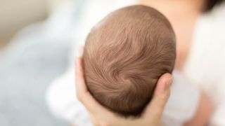 A baby&#039;s head, with some hair on top, features prominently at center. A hand holds the head up. An adult in the background is holding the baby.