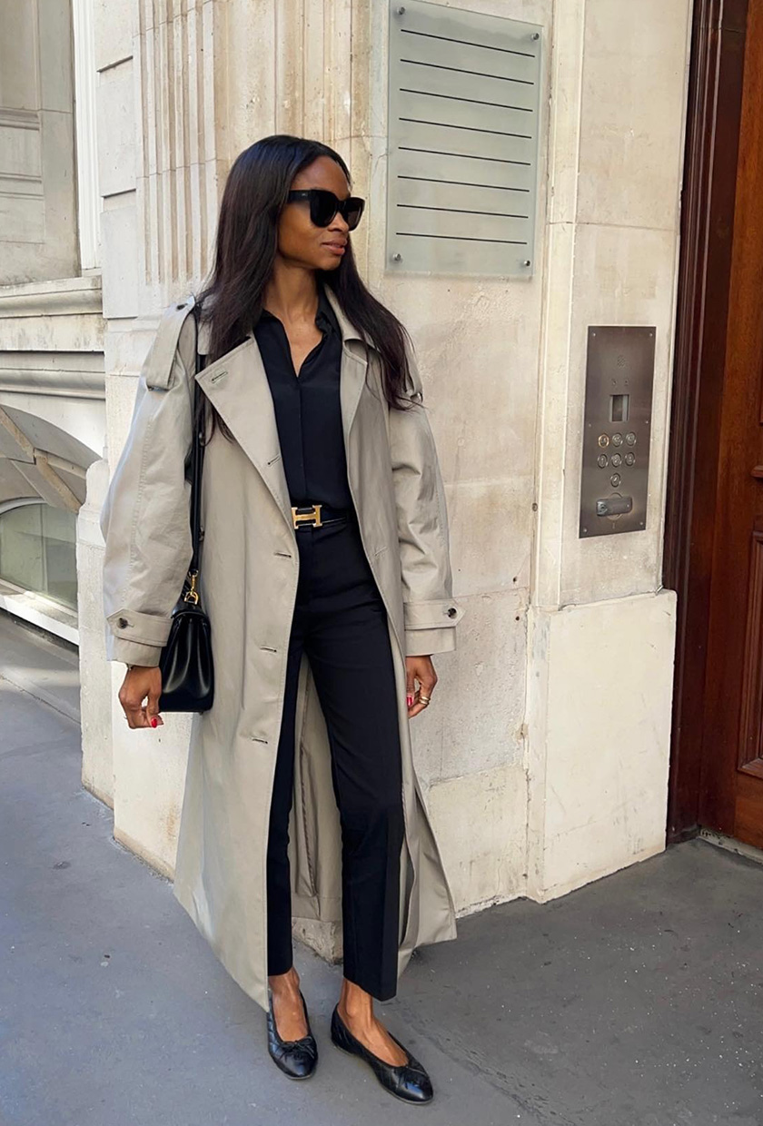 A woman standing in a doorway wearing a tan trench coat styled with a black button-down shirt, black tailored trousers, black ballet flats, a black belt, black sunglasses, and a black shoulder bag.