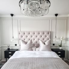 A white bedroom with a large padded headboard, black bedside tables and a large glass chandelier