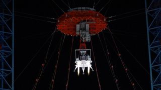 A lander with four legs, uses a red parachute to drop to the surface during a nighttime test. 