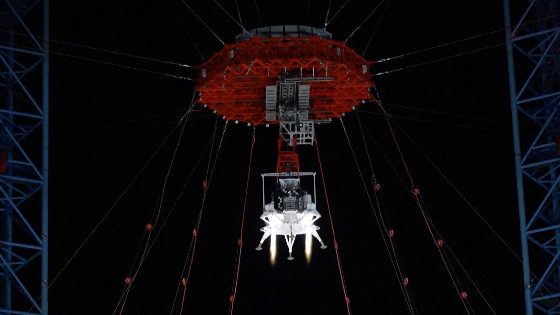 A lander with four legs, uses a red parachute to drop to the surface during a nighttime test. 