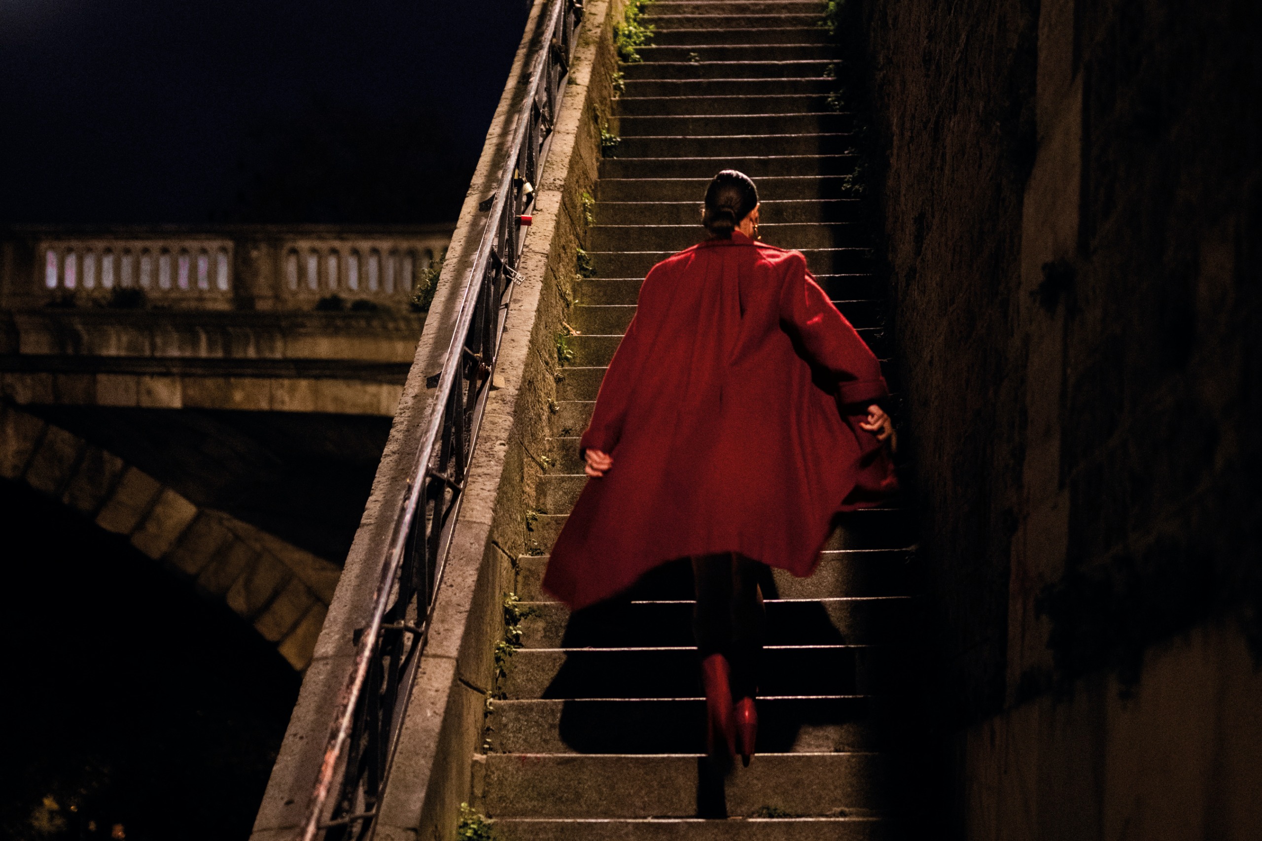 A woman in a flowing red coat climbs a stone staircase at night towards an arched stone bridge, her coat caught mid-swing.