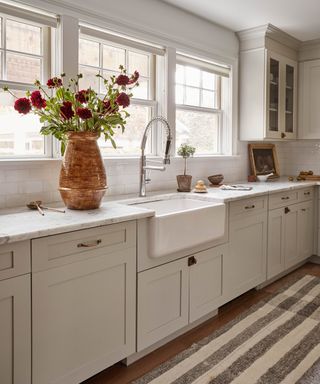 Pale gray kitchen cabinetry with a butler sink below a row of windows.